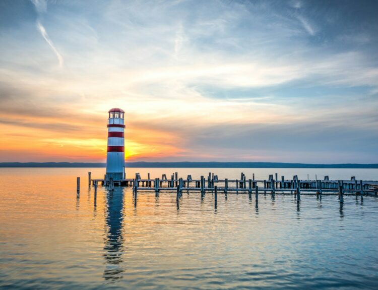 photo of white and red lighthouse beside wooden beach dock under clear blue sky